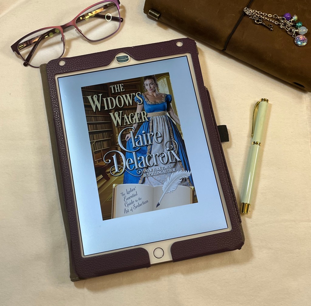 iPad showing cover of The Widow's Wager by Claire Delacroix on white background with pen, reading glasses, and a notebook in the background.