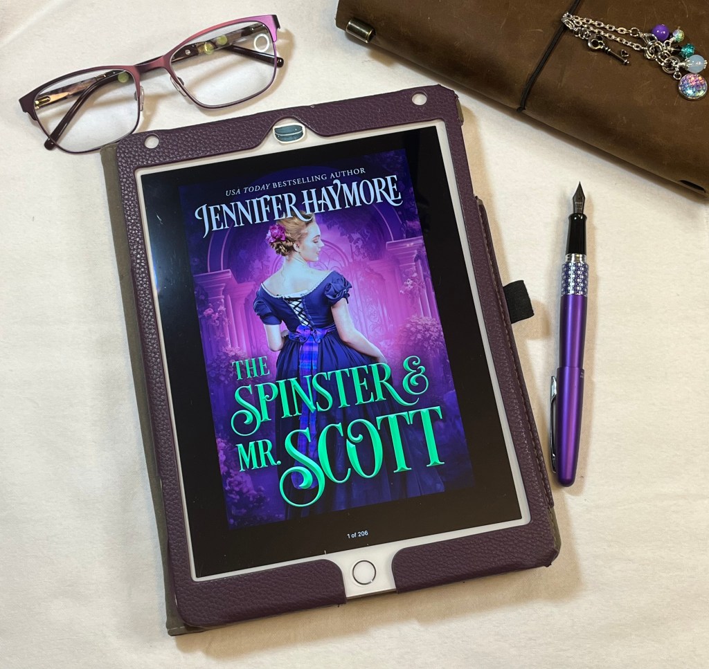 iPad showing cover of The Spinster and Mr. Scott with fountain pen, reading glasses, and notebook in the background