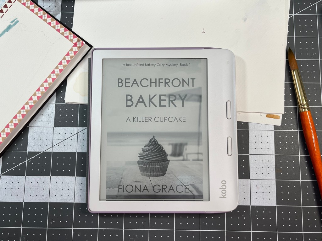 Kobo Libra 2 showing cover of Beachfront Bakery: A Killer Cupcake by Fiona Grace with paintbrush and paper in the background.