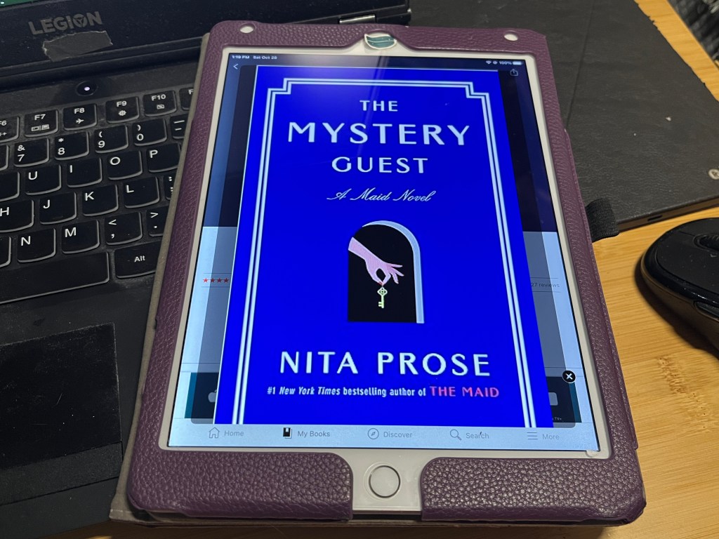 iPad showing cover of book The Mystery Guest by Nita Prose with a laptop keyboard, mouse, and ereader in the background.